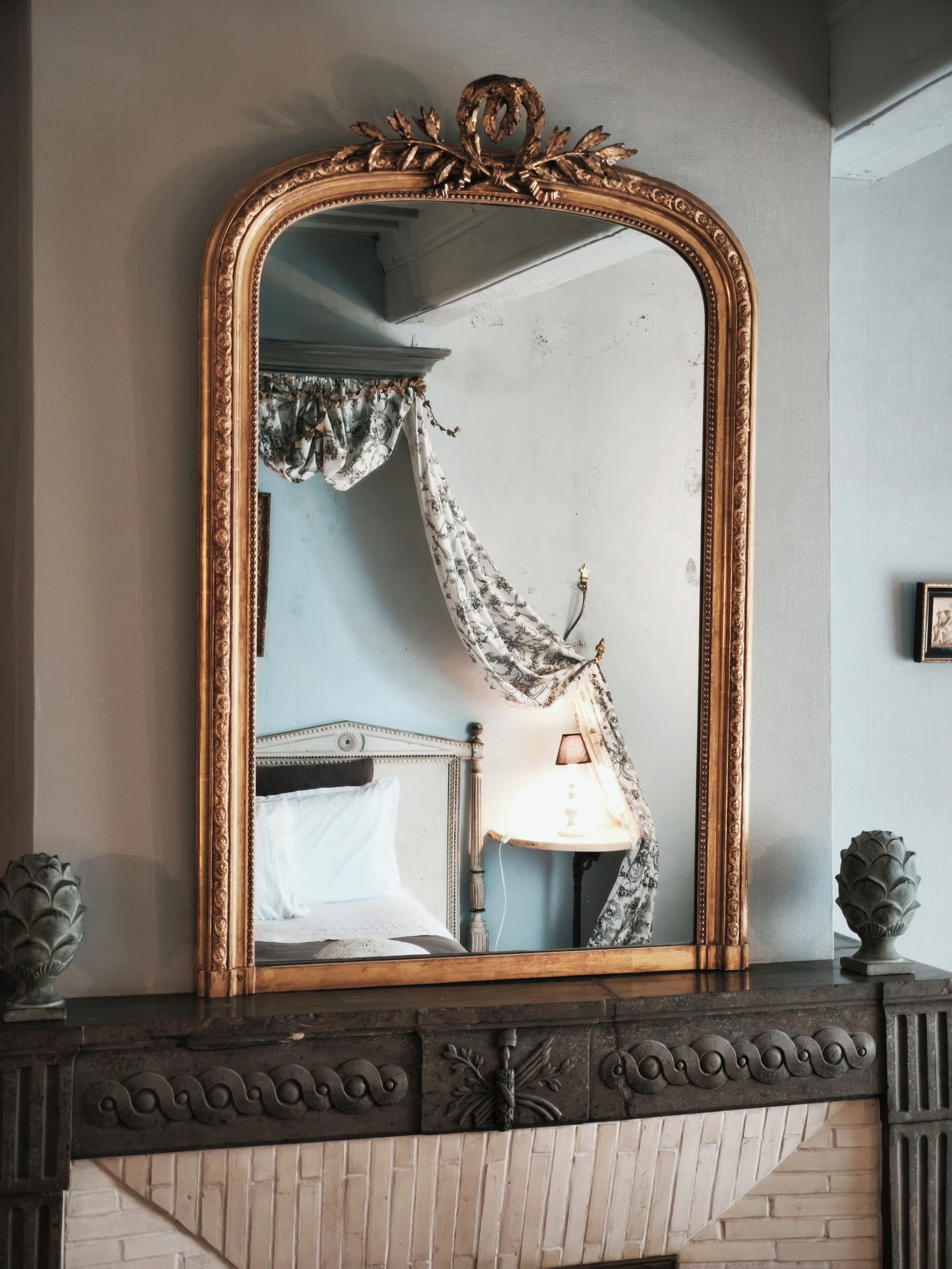 A room with a large ornate mirror, a bed with white bedding, a nightstand with a lamp, and a decorative fireplace mantel.