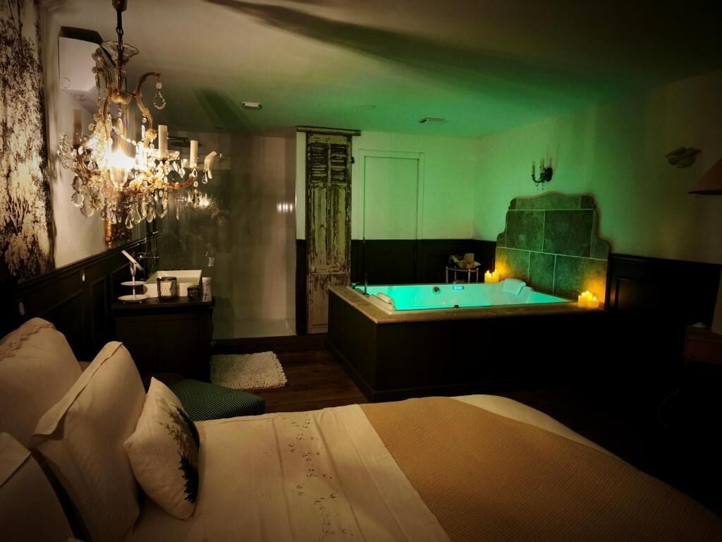 A dimly lit room with a large bed, a sitting area with a chair and a small table, a bathtub, and a chandelier.