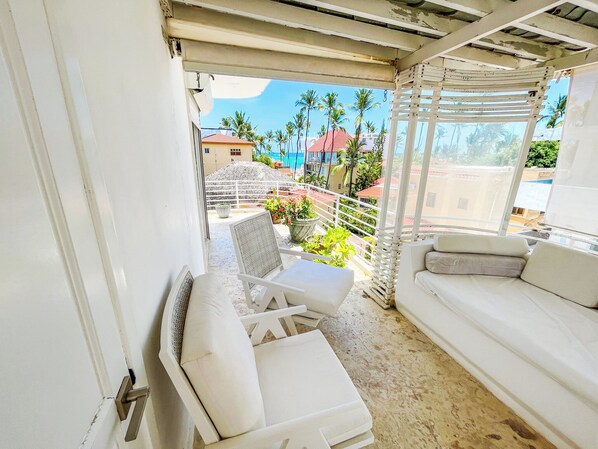 A balcony with a white sofa, a wicker chair, and a table. There are potted plants and a view of palm trees and buildings in the background.
