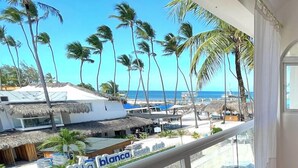 Uma varanda com vista para uma praia, palmeiras e uma placa para 'La Blanca Beach Club'.