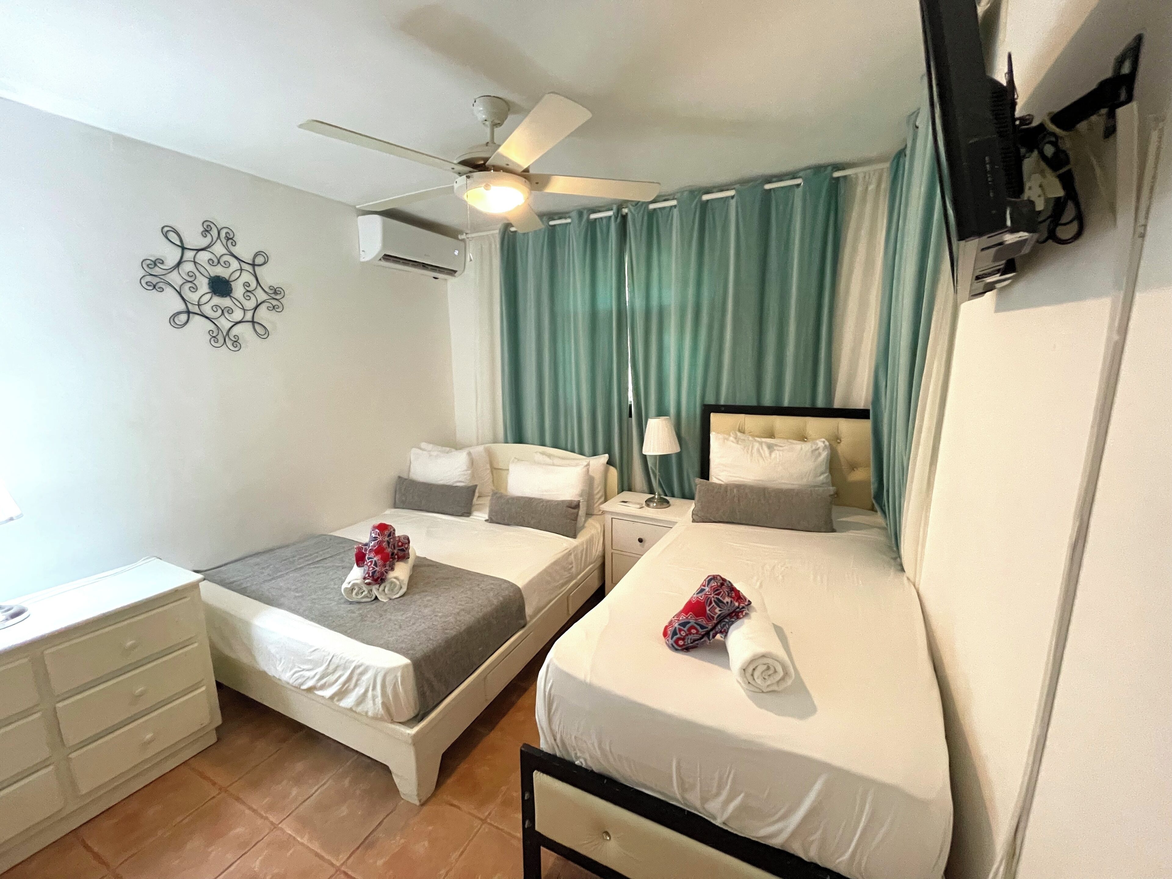 Quarto de hotel com duas camas, um ventilador de teto, ar condicionado, uma escrivaninha e uma televisão.