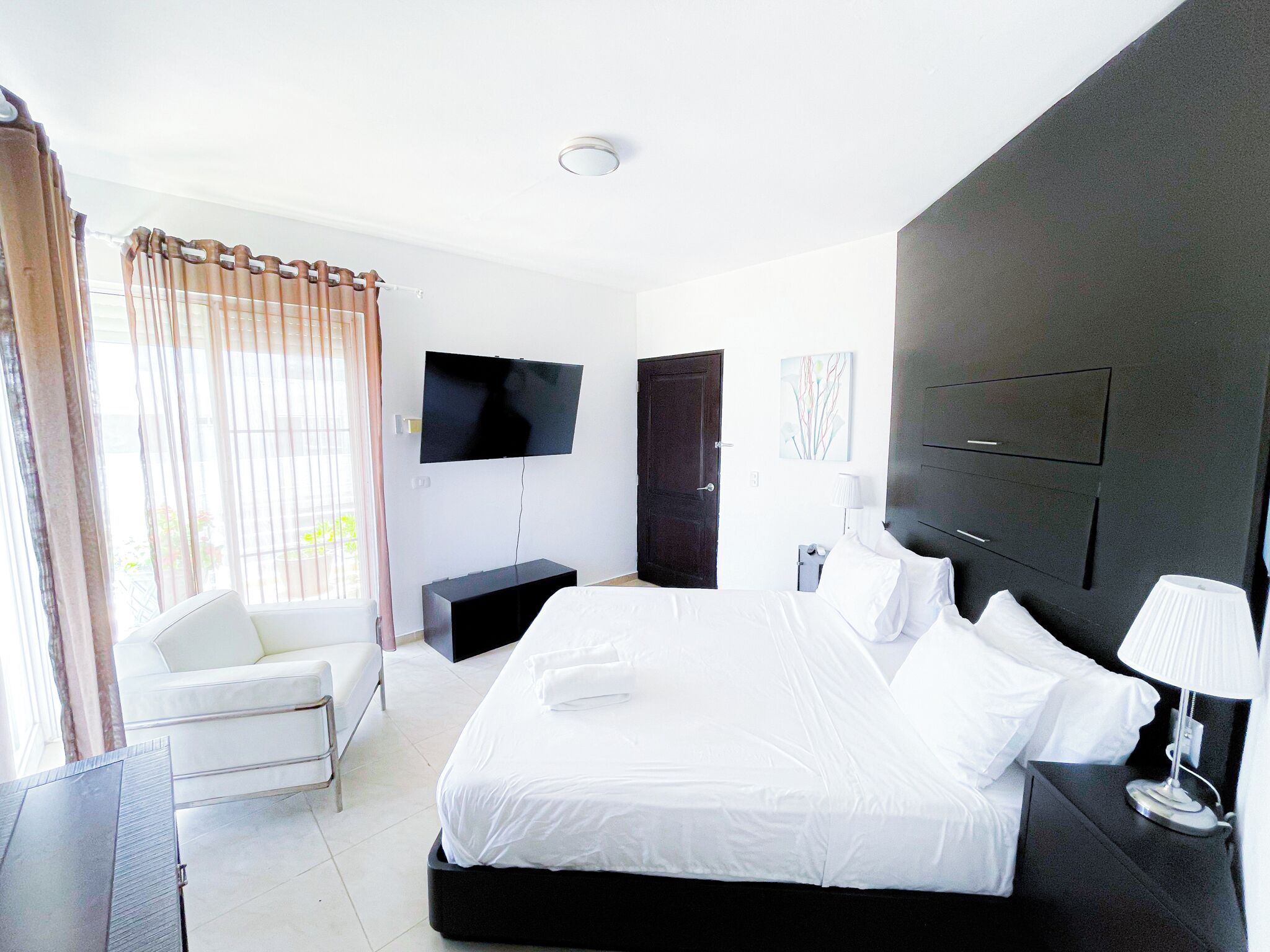 A modern hotel room with a large bed, a flat-screen TV mounted on the wall, a bedside table with a lamp, and a window with curtains.