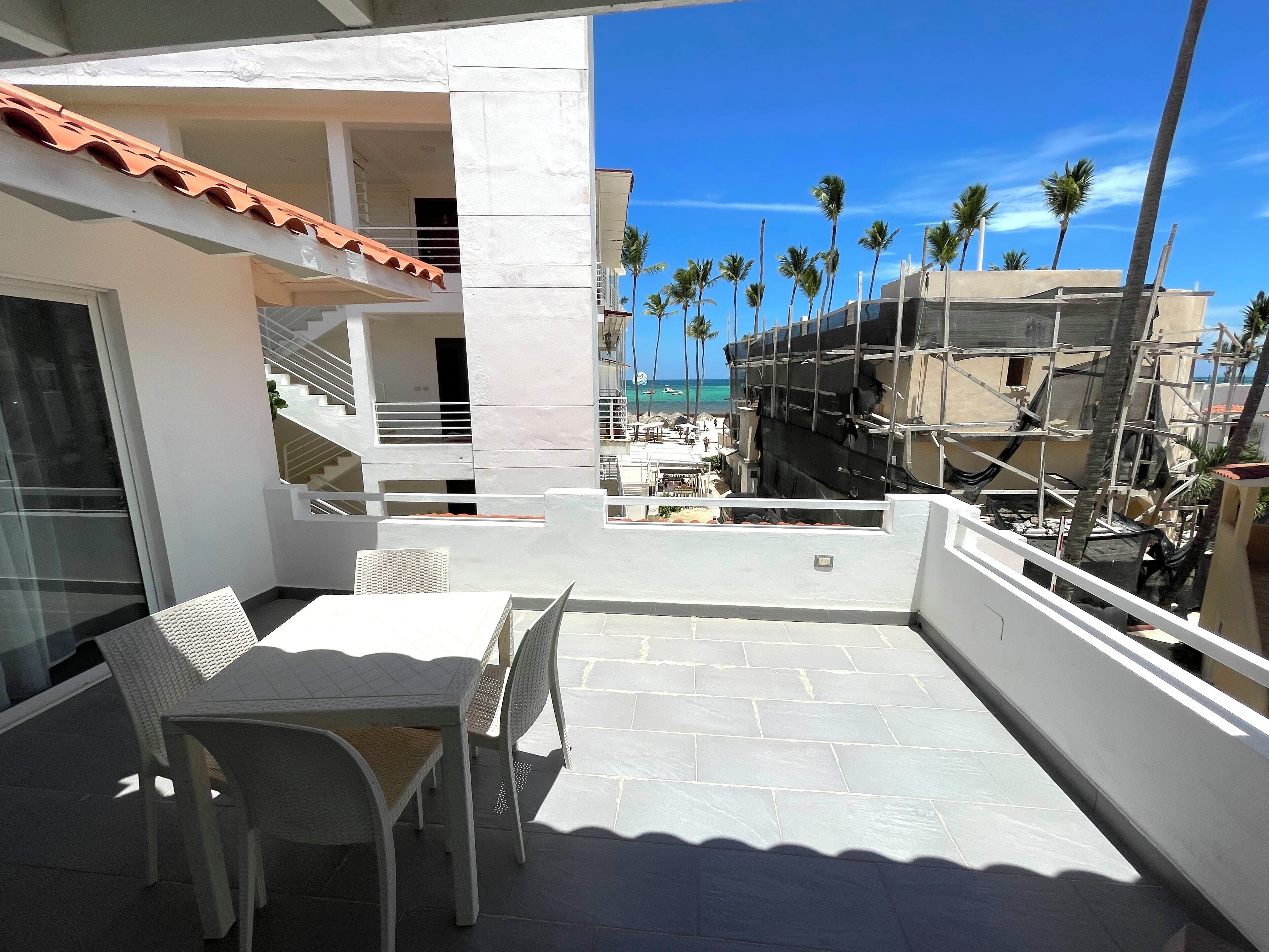 A balcony with a table and chairs, a tiled floor, and a view of palm trees and a beach.