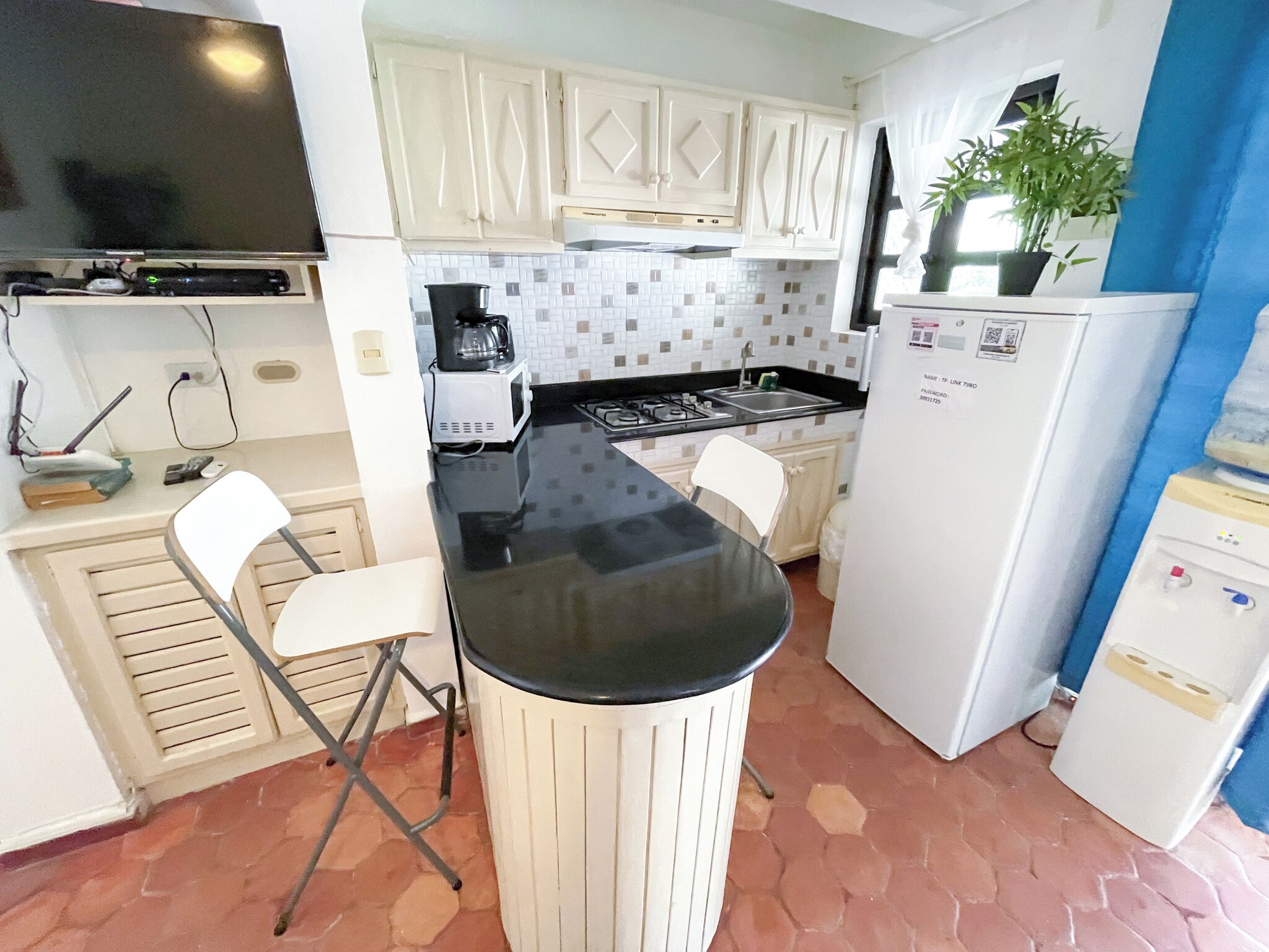 Una cocina compacta con armarios blancos, una encimera negra, un refrigerador blanco y un taburete pequeño.