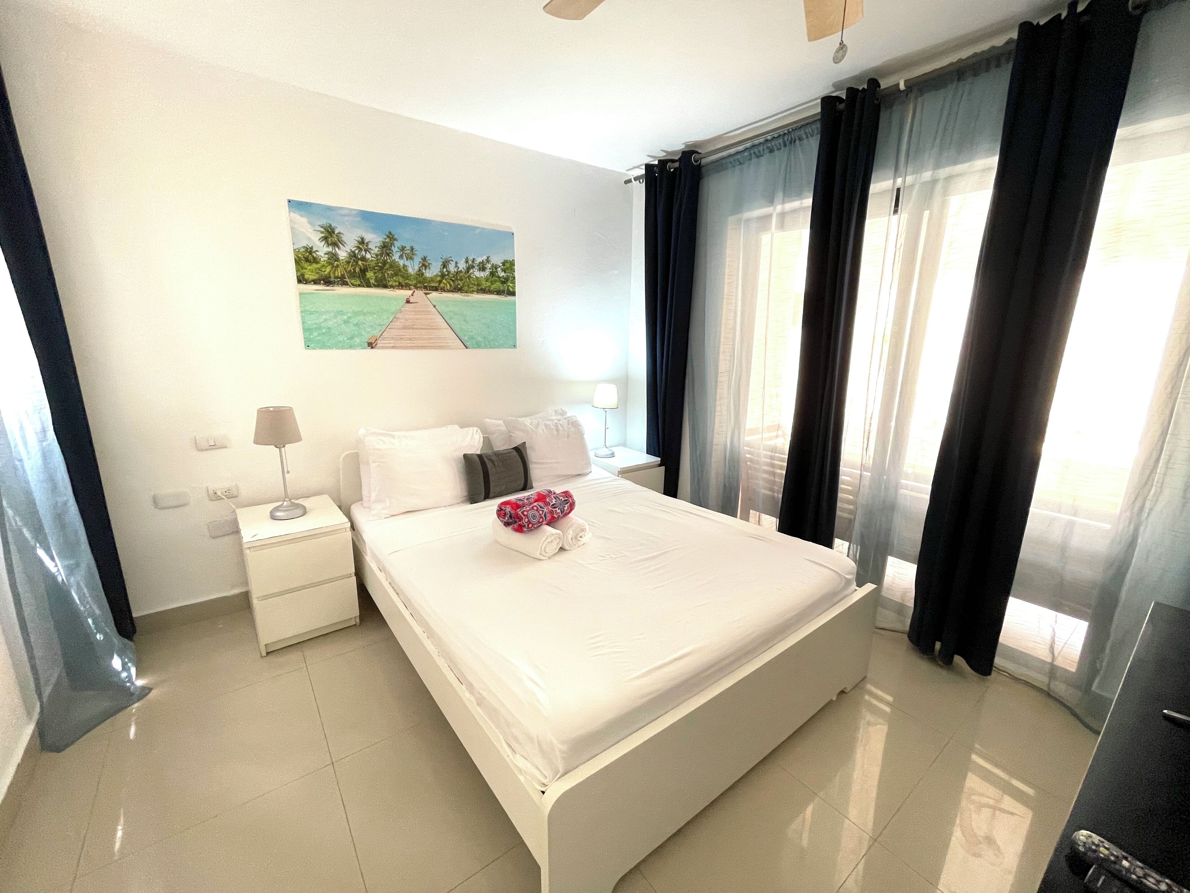 Hotelkamer met een groot bed, nachtkastje, lamp en een schilderij van een strand.