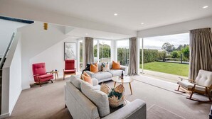 A spacious living room with a grey sofa set, a red armchair, and a white rocking chair. A staircase is visible on the left, leading to an upper level. - 7th Heaven (Napier)