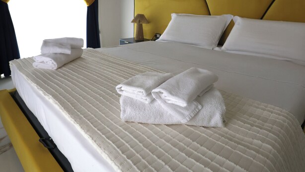 Een net opgemaakt bed met witte lakens en handdoeken, een geel hoofdeinde en een nachtkastje met een lamp.