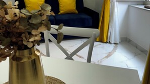 Een woonkamer met een blauwe bank, gele kussens, een witte tafel en een gouden vaas met gedroogde bloemen.