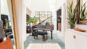 Un piano de cola imponente en una habitación amplia con techo alto y ventanas grandes, una planta en maceta y un aparador con cajones.