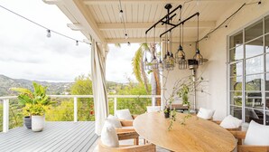 Una terraza de madera con una mesa redonda y sillas, plantas en macetas y lámparas colgantes.
