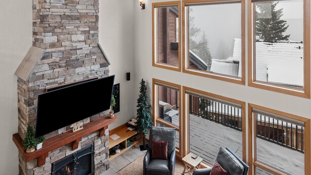 A spacious living room with a stone fireplace, leather sofas, and large windows offering a view of the snowy outdoors. - Northwest Passage Retreat Ski-In/Ski-Out + Hot Tub (Sandpoint)