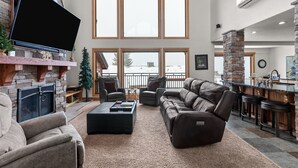 A spacious living room with a fireplace, leather sofas, a flat-screen TV, and large windows. - Northwest Passage Retreat Ski-In/Ski-Out + Hot Tub (Sandpoint)