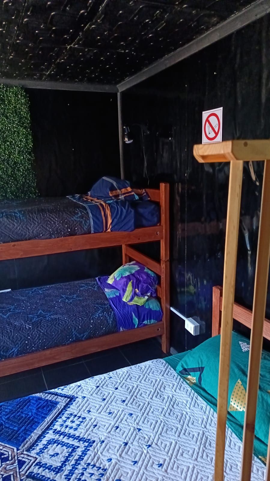 Una cama individual con un edredón estampado en azul y morado, una almohada verde y amarilla, y un letrero de prohibido fumar.
