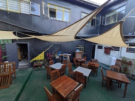 Un área de estar al aire libre con mesas y sillas, protegida por grandes toldos, y una escalera que conduce a un nivel superior.