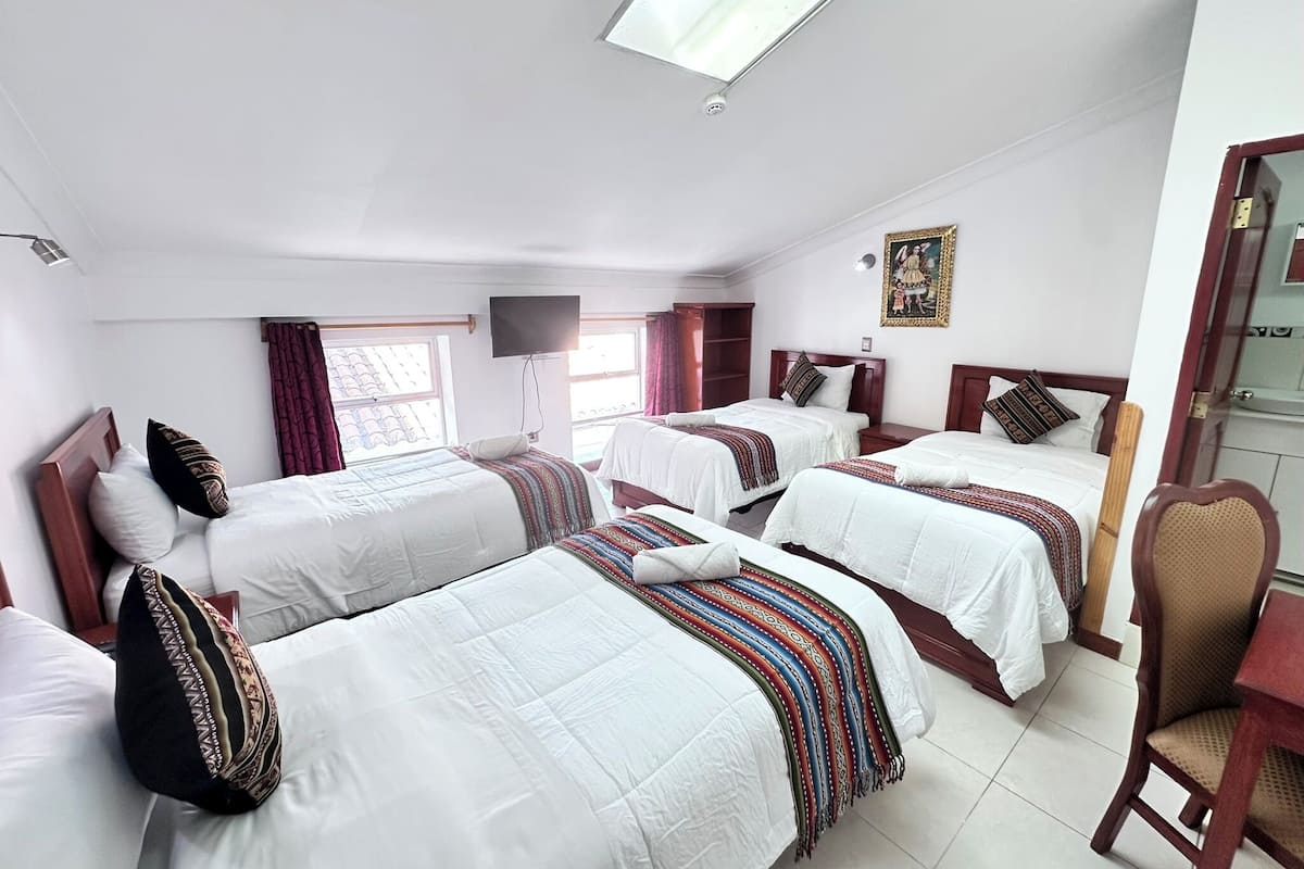 A hotel room with three single beds, a TV mounted on the wall, a window with curtains, and a small table with a chair.