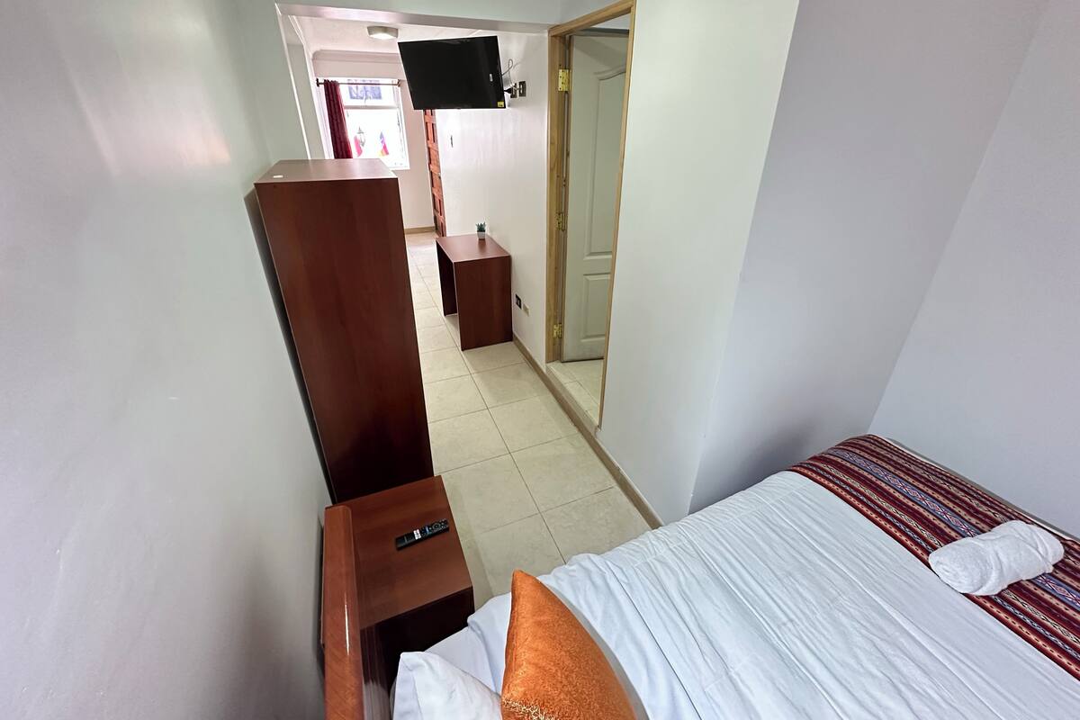 A hotel room with a bed, a wooden nightstand, a TV mounted on the wall, and a door leading to another room.