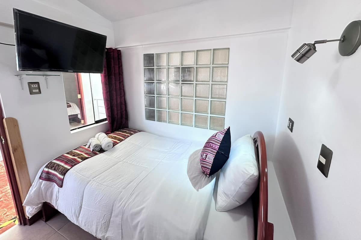 A neatly arranged bedroom with a bed, a television mounted on the wall, a window with a view, and a wall-mounted lamp.