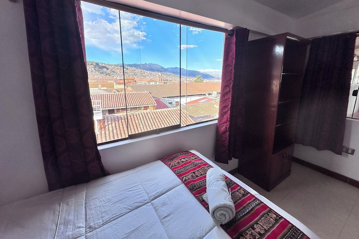 A hotel room with a bed, a window with curtains, and a view of a cityscape with mountains.