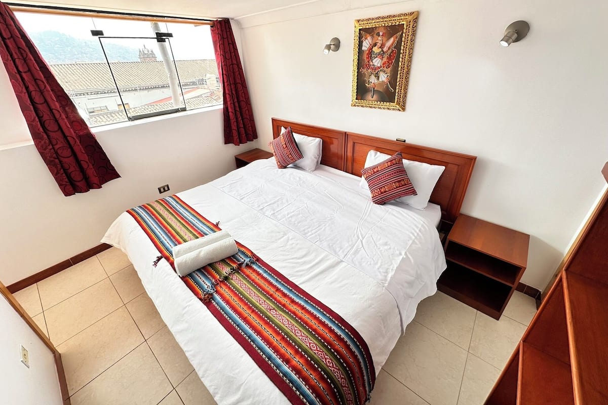A hotel room with a large bed, a window with curtains, a wall-mounted picture, and a small wooden nightstand.