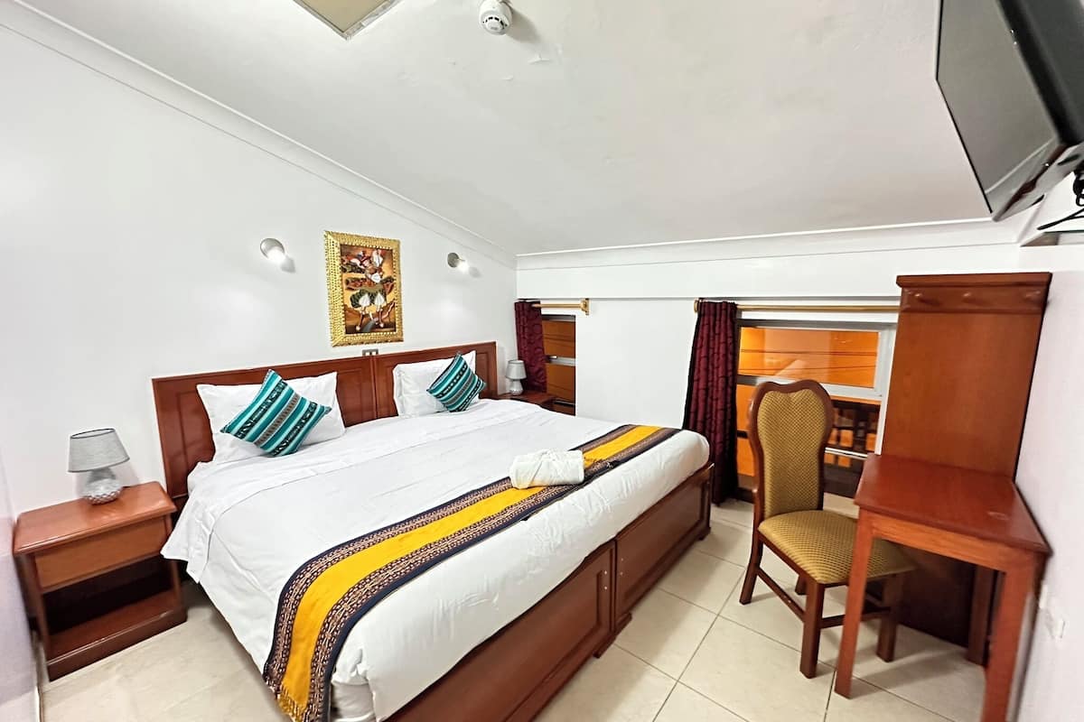 A hotel room with a bed, bedside table, lamp, desk, chair, and a television.