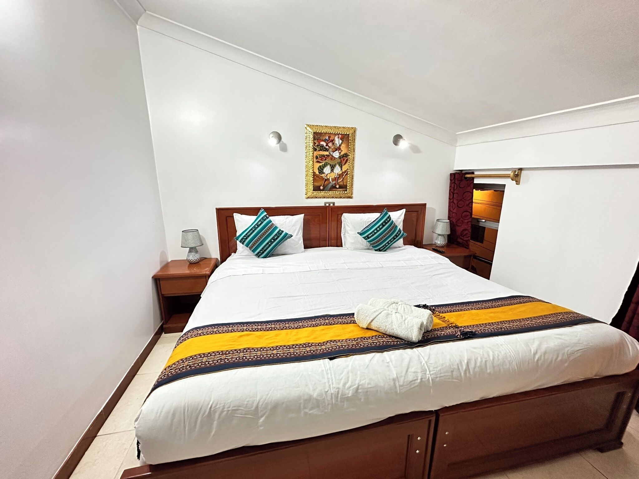 A neatly made bed with a patterned bedspread, two pillows, a nightstand with a lamp, and a wall-mounted painting.