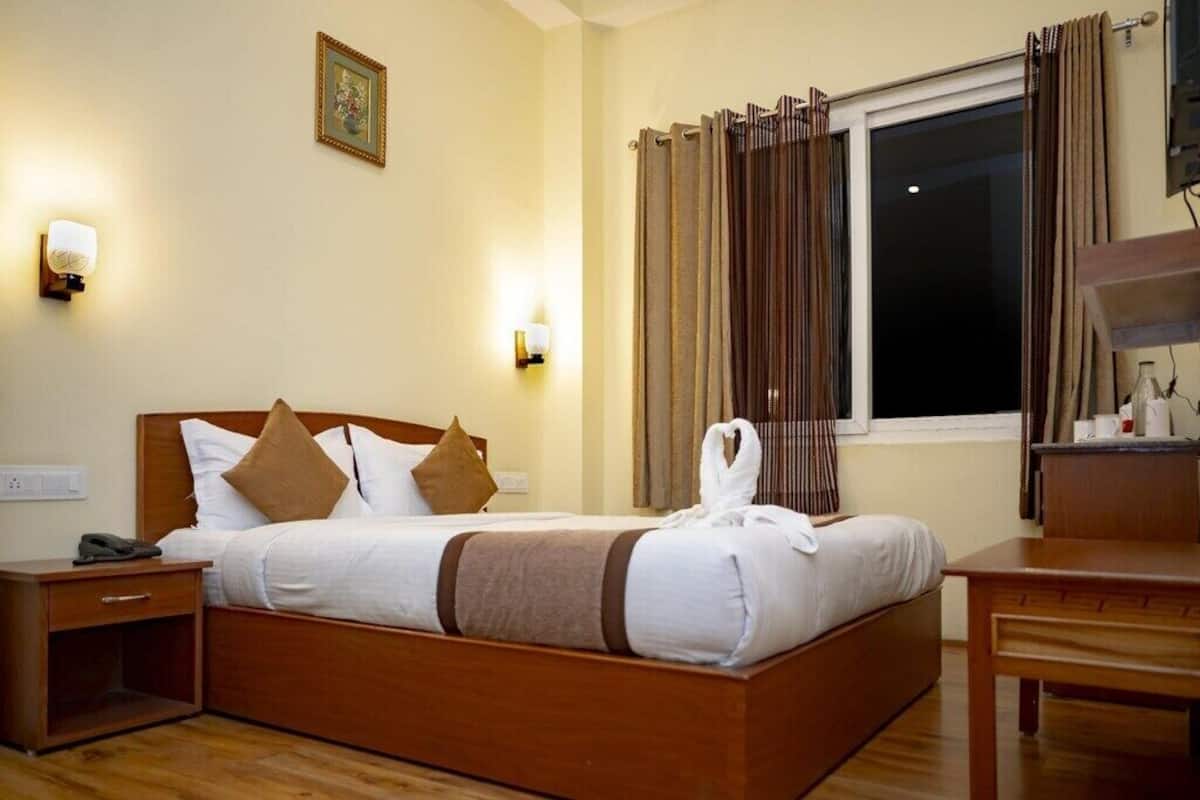 A hotel room with a wooden bed, two bedside tables, a nightstand, a window with curtains, and wall-mounted lights.