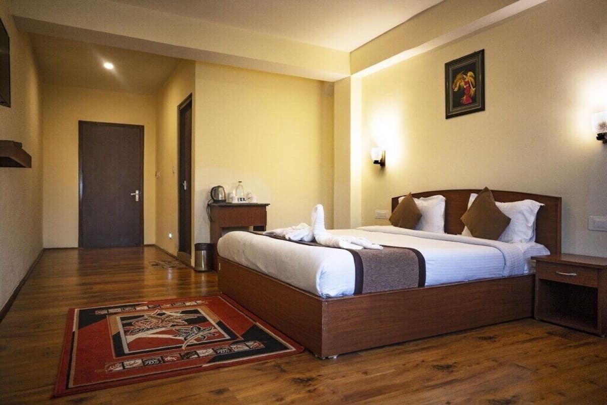 A hotel room with a wooden bed, a nightstand, a small table with a lamp, and a door in the background.