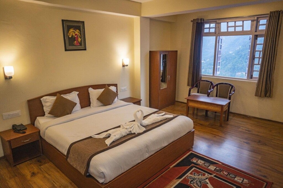 A hotel room with a large bed, bedside tables, a desk, a chair, a window with curtains, and a framed picture on the wall.