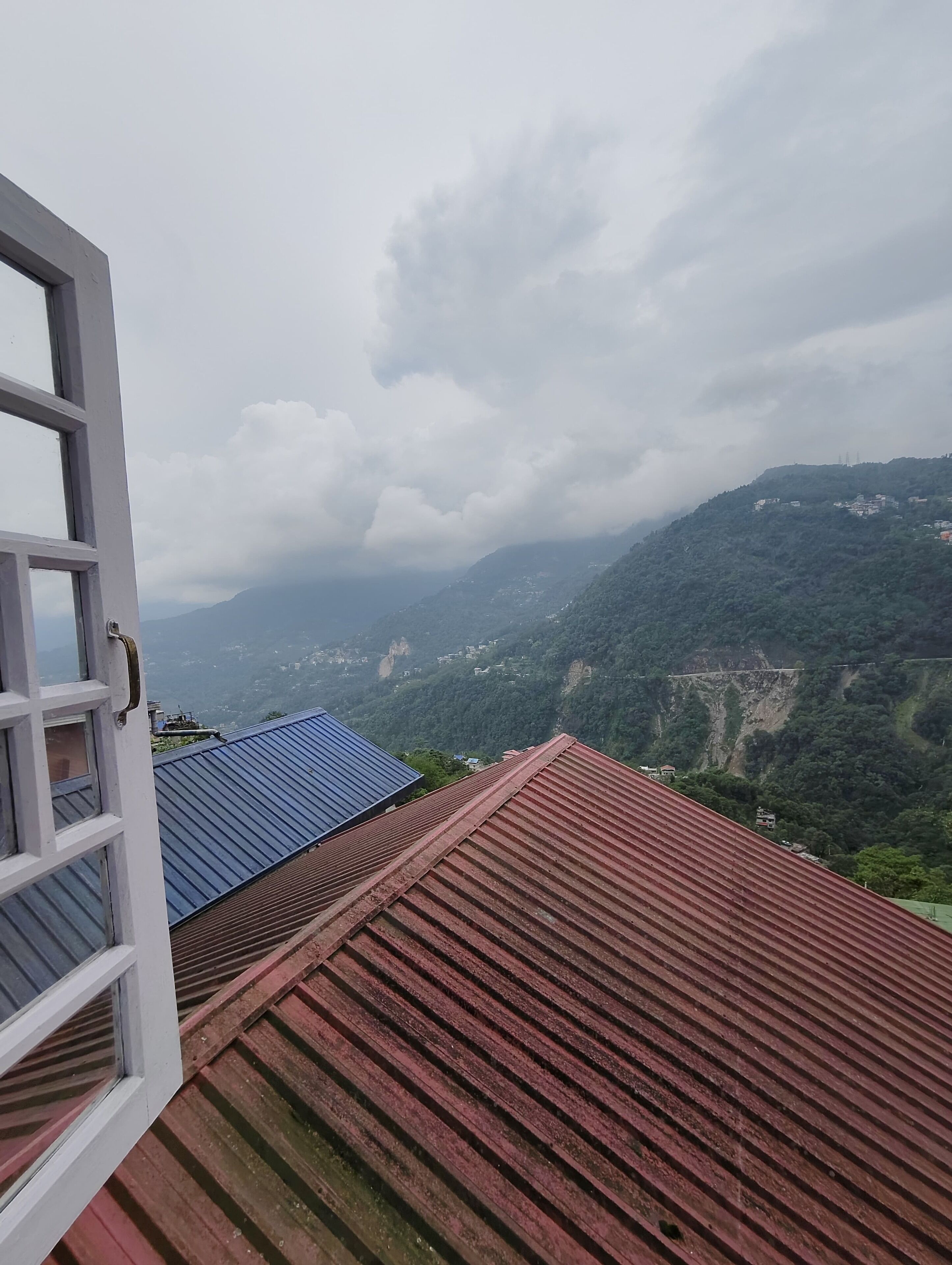 A view from a window overlooking a mountainous landscape with a red-roofed building and a blue-roofed structure in the distance.
