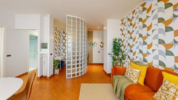 Ein modernes Wohnzimmer mit einer gebogenen Trennwand, einem Sofa mit gemusterten Kissen und einer Wand mit geometrischen Mustern.
