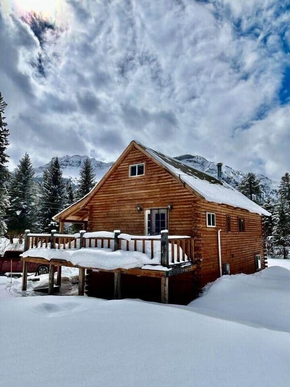 A wooden cabin with a porch, surrounded by snow-covered ground and trees, with mountains in the background.