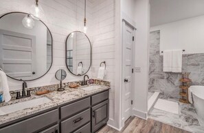 A modern bathroom with two sinks, a large mirror, and a marble shower area.