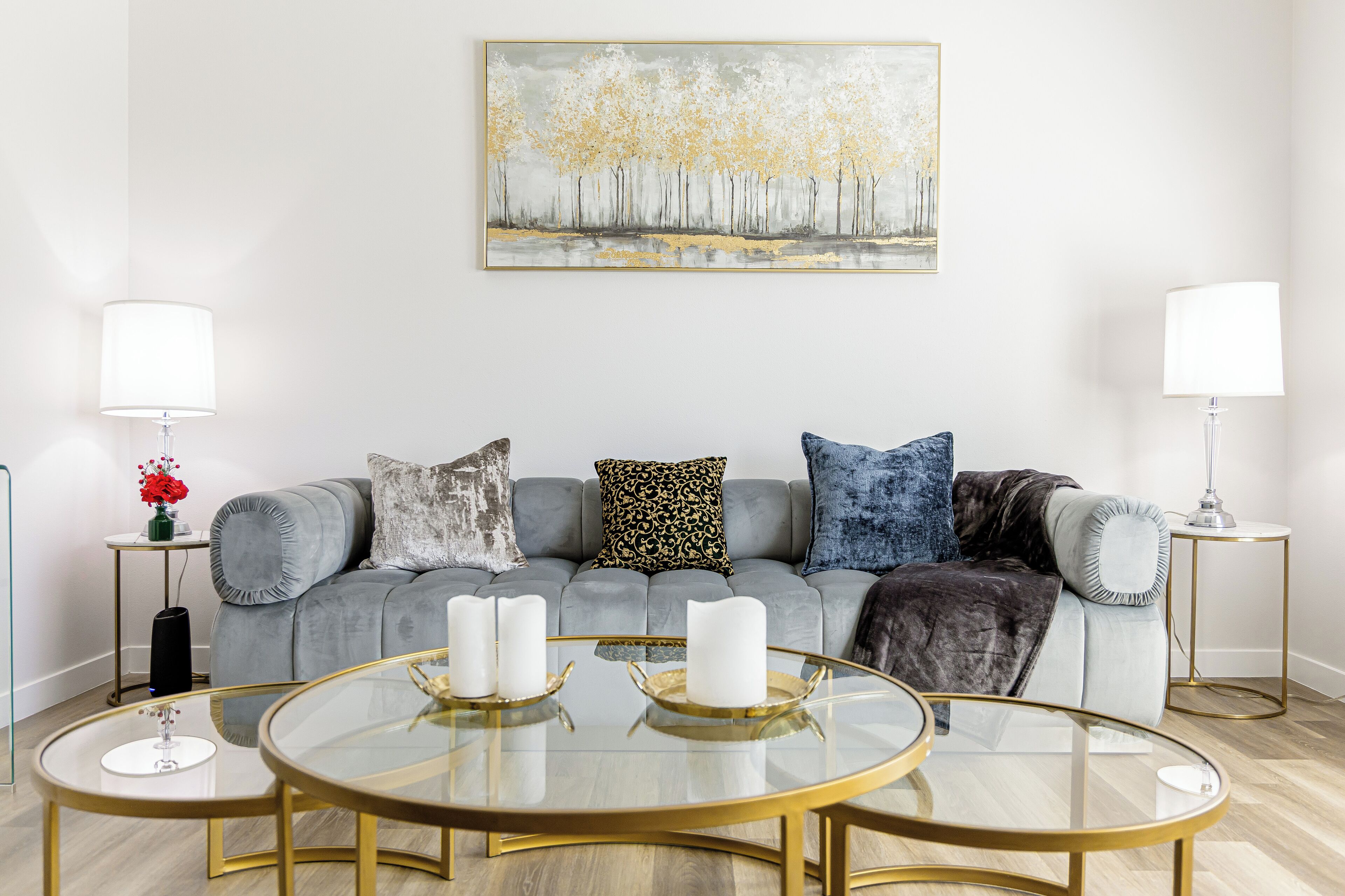 Un salon moderne avec un canapé gris, trois petites tables rondes en verre dotées de cadres dorés et deux tables d’appoint avec des lampes.