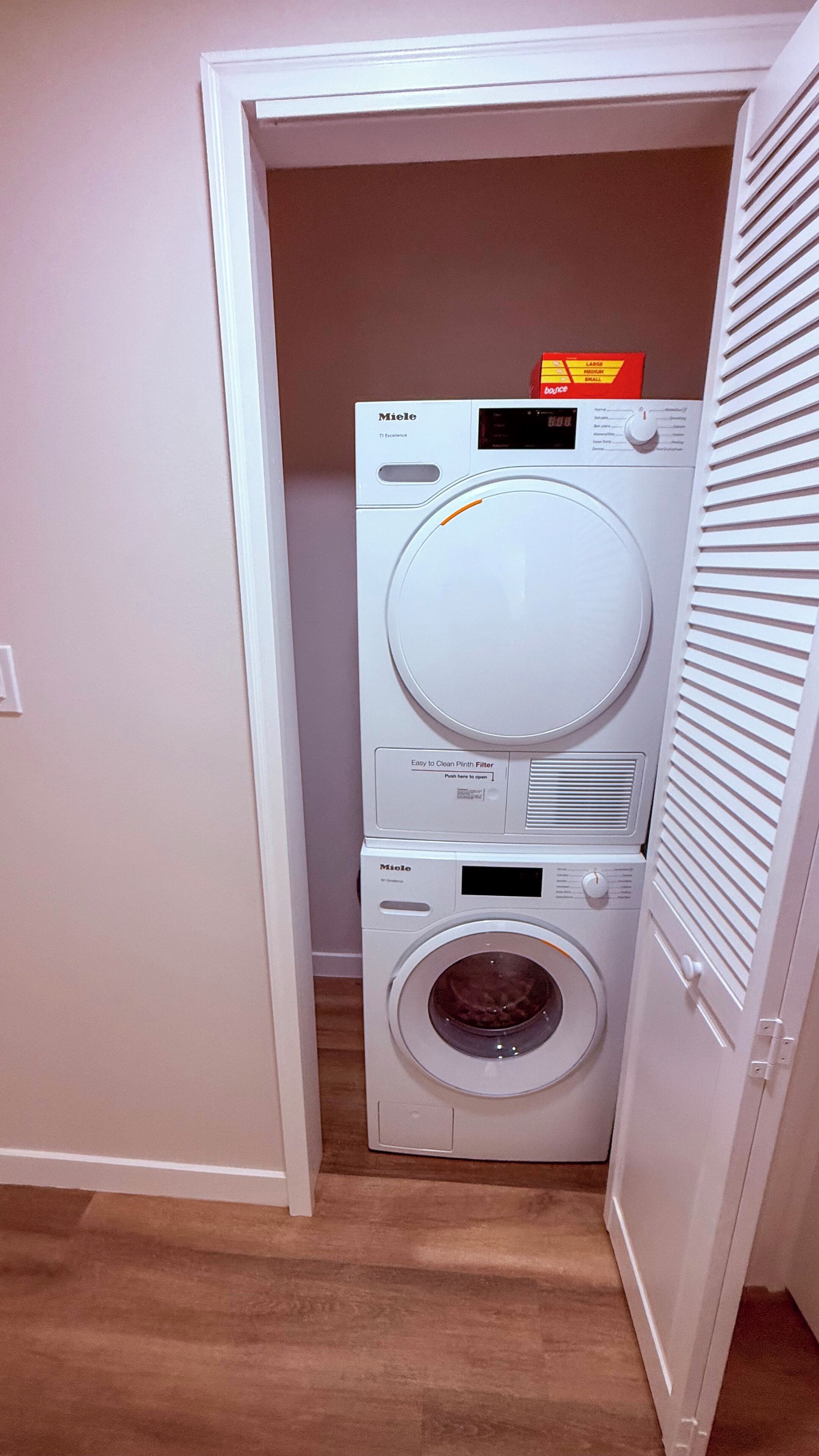 Lavadora y secadora apiladas, de color blanco y tamaño compacto, en un pequeño cuarto de lavado con piso de madera.