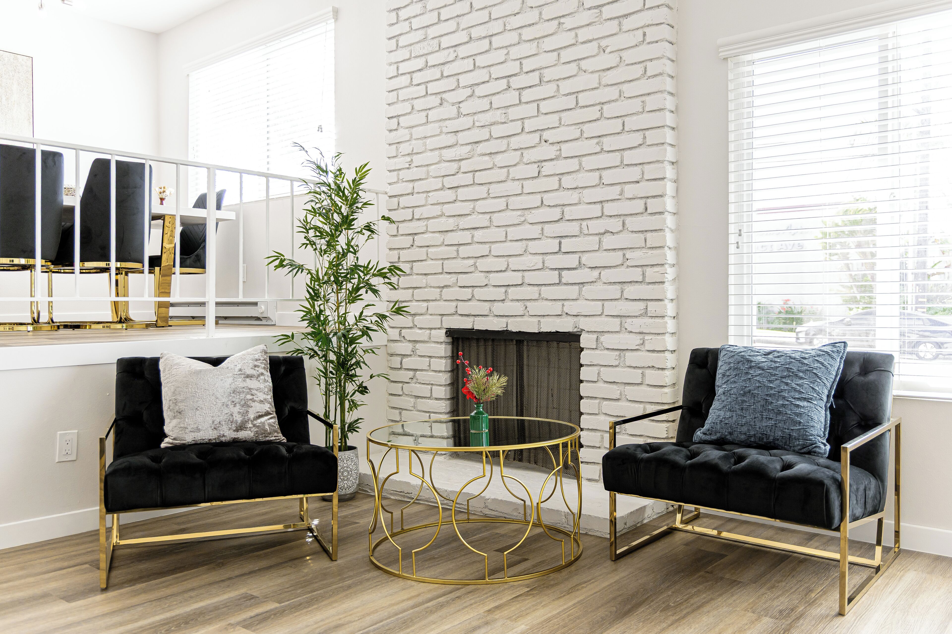 Un salón moderno con dos sillones tapizados de color negro, una mesa de centro con marco dorado y una chimenea de ladrillo blanco.