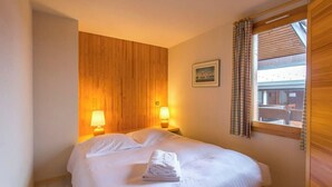 A hotel room with a wooden headboard, two bedside lamps, a neatly made bed with a folded towel, a framed picture on the wall, and a window with curtains. - Creux de l'ours D172 - 9 guests - Ski in - Ski out (Méribel)