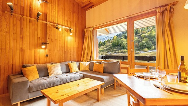 A cozy wooden interior with a sofa, table, and large windows offering a view of a mountainous landscape. - Creux de l'ours D172 - 9 guests - Ski in - Ski out (Méribel)