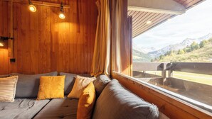 A cozy room with a wooden interior, a sofa with cushions, and a large window offering a mountain view. - Creux de l'ours D172 - 9 guests - Ski in - Ski out (Méribel)