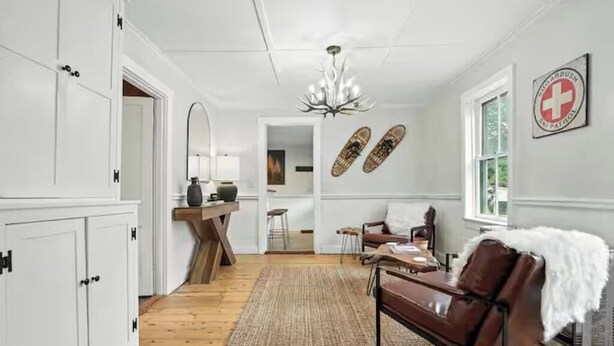 A spacious living area with a brown leather sofa, a wooden coffee table, and a chandelier. There is a white cabinet on the left and a window on the right.