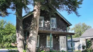 A two-story wooden house with a porch, surrounded by trees and greenery.