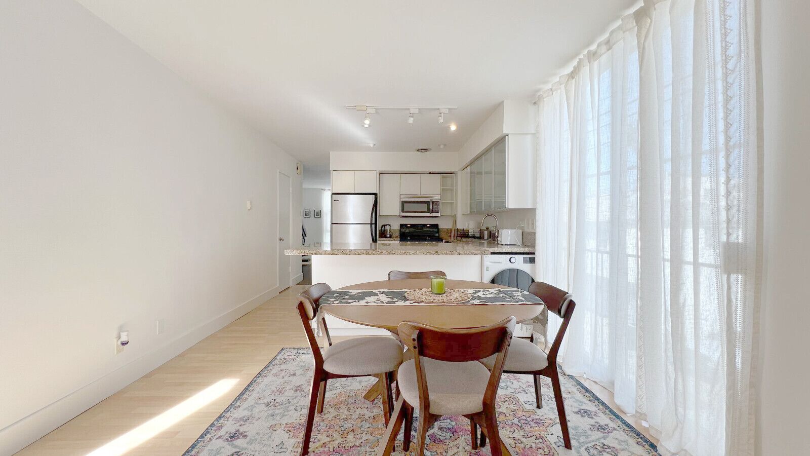 Une cuisine moderne avec des meubles blancs, une table à manger avec des chaises, et une vue sur la cuisine à travers une fenêtre.