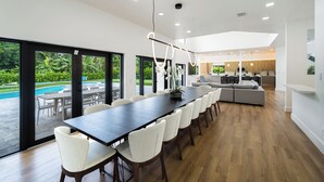 A modern kitchen with a central island, a dining area with a long table and chairs, and a view of a swimming pool. - Villa Rio Grande 8BR Lazy River Pool + Pickleball (Miami)