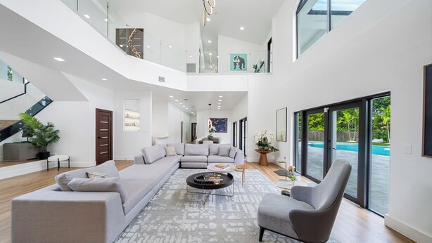 A spacious living area with a large sectional sofa, a round coffee table, and a grey armchair. A staircase leads to an upper level with a glass railing. - Villa Rio Grande 8BR Lazy River Pool + Pickleball (Miami)