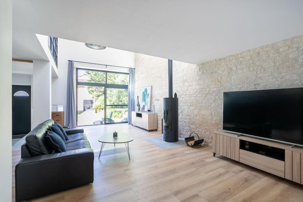 A modern living room with a black leather sofa, a flat-screen TV, a stone wall, and a large window.
