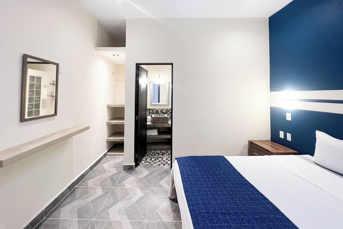 A hotel room with a bed, a nightstand, a mirror, and a bathroom visible through an open door.