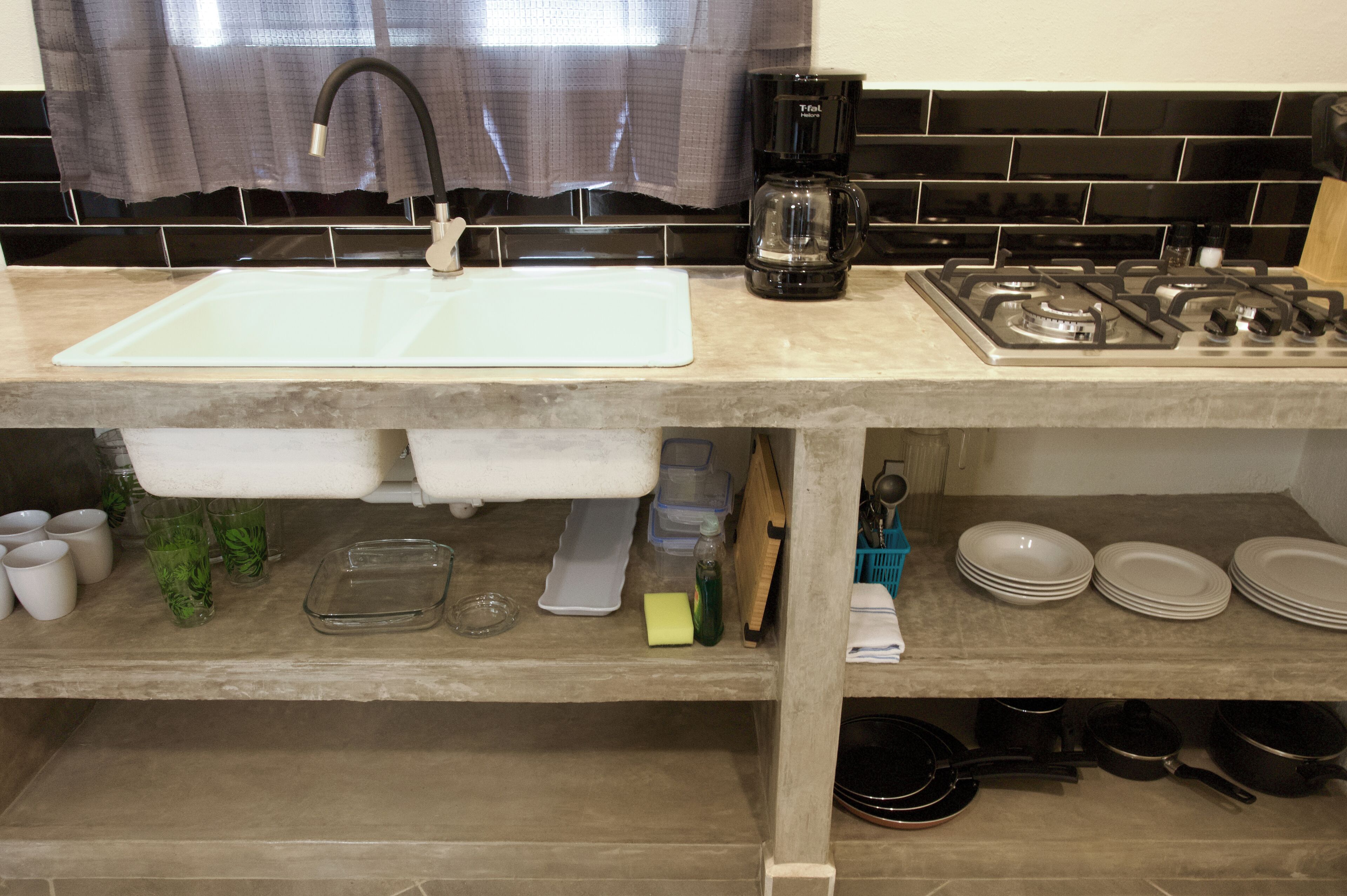 Una cocina con un lavabo blanco, cafetera negra y estufa. Hay tazas, un escurreplatos y una tabla de cortar en la encimera.