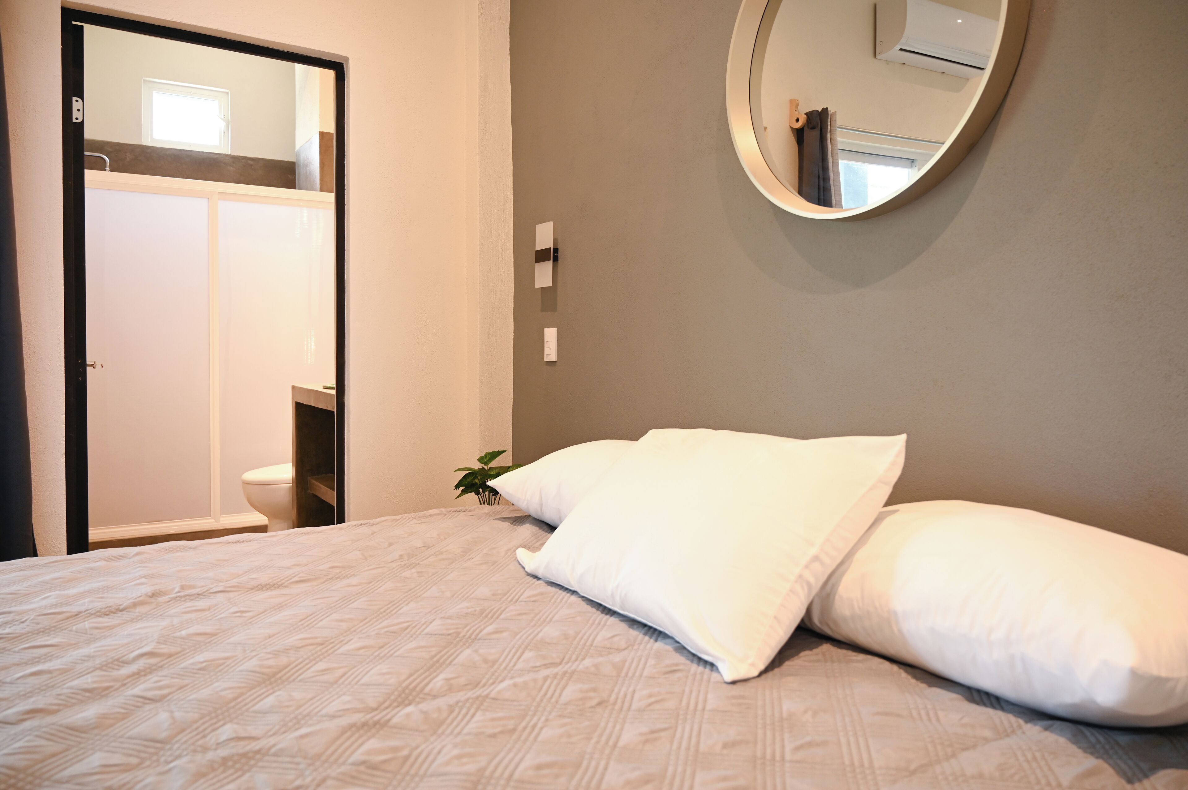 Een slaapkamer met een bed, kussens, een ronde spiegel en uitzicht op een badkamer met toilet en wastafel.