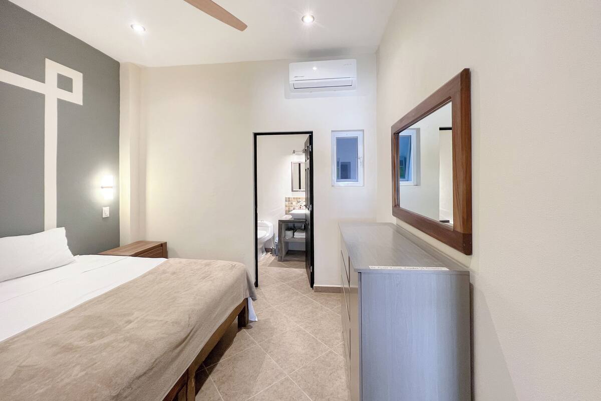 A bedroom with a bed, a ceiling fan, an air conditioning unit, a dresser, and a mirror.