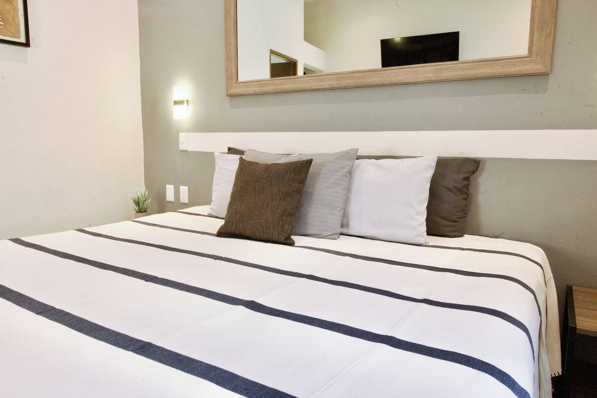 A neatly made bed with striped bedding and a headboard, a small potted plant, and a wall-mounted mirror.