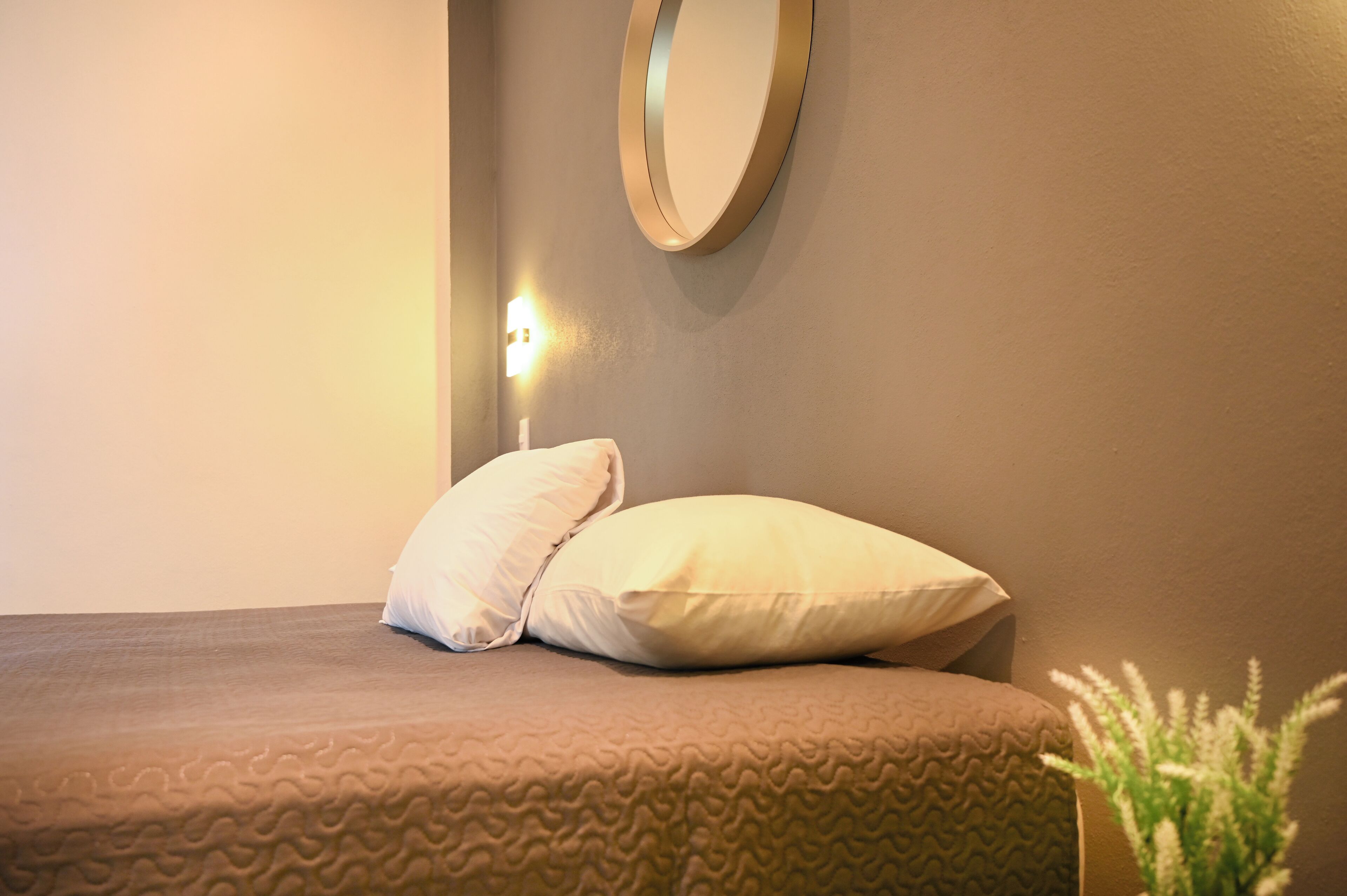 A neatly made bed with two pillows, a round mirror on the wall, and a small wall-mounted light.